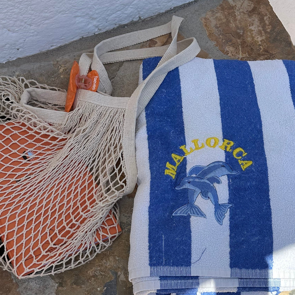 Beach scene featuring a striped Mallorca towel and a net bag containing Faeves products, capturing the essence of a relaxed summer day.