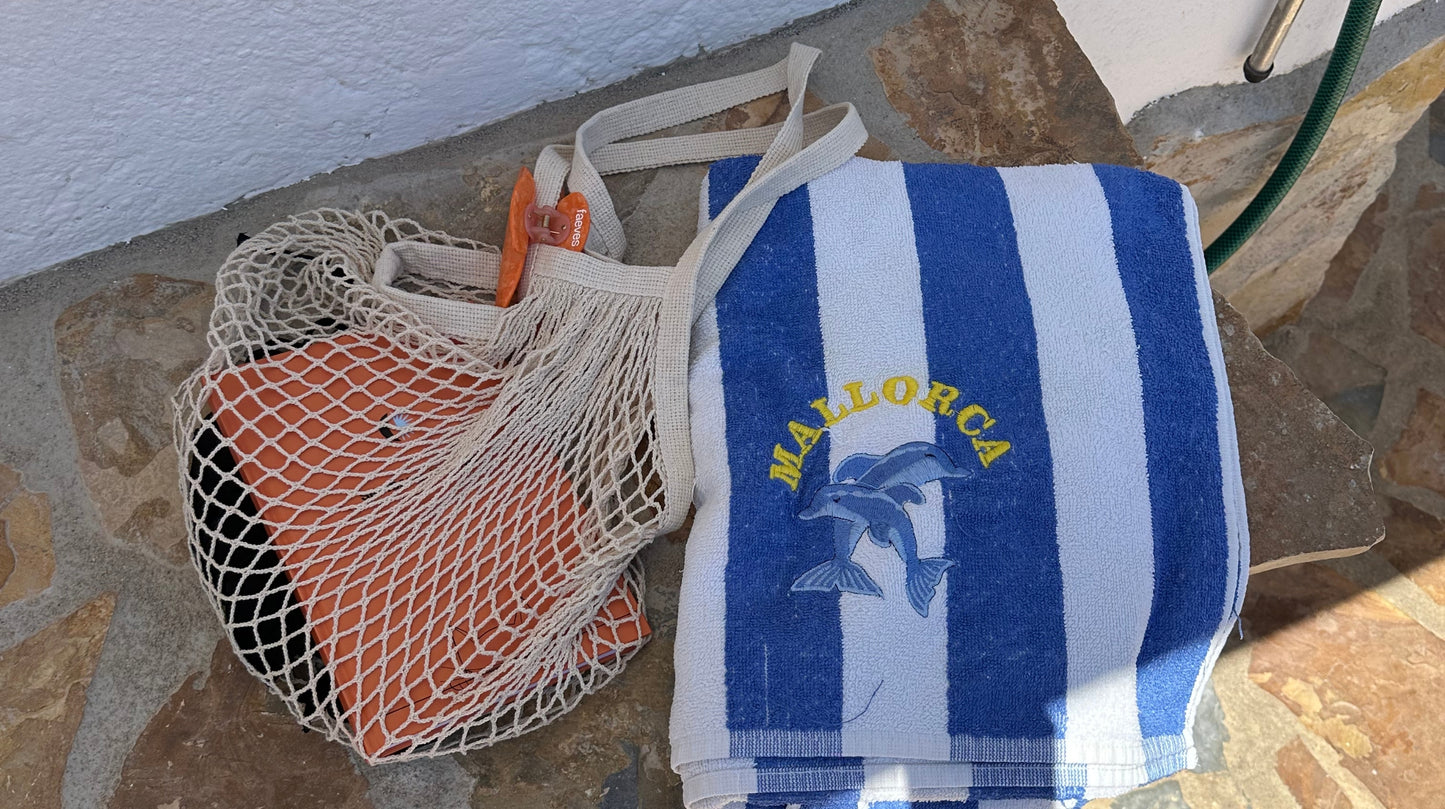 Beach scene featuring a striped Mallorca towel and a net bag containing Faeves products, capturing the essence of a relaxed summer day.